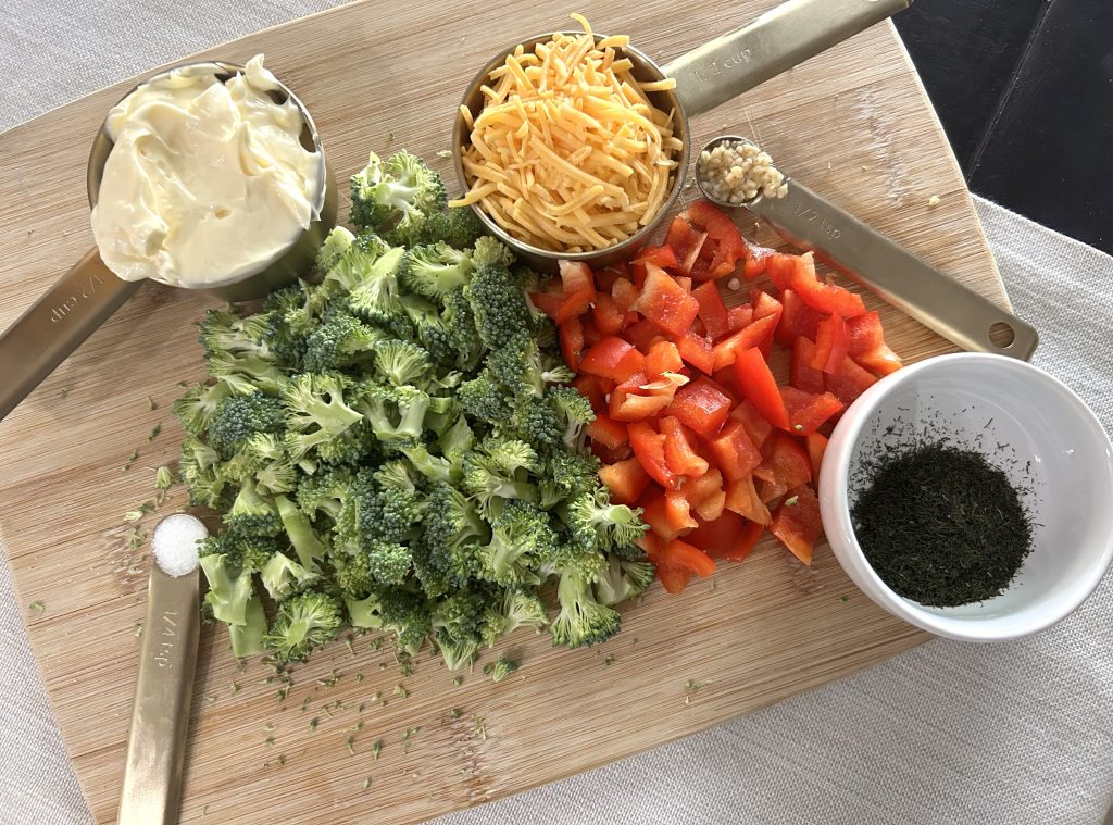 A wooden cutting board with prepared ingrdients of chopped broccoli, red pepper, mayo, shreeded cheedar cheese, and dill