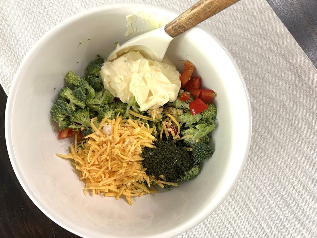 A white mixing bowl on a gray table runner of chopped ingredients with a wooden spatula ready to be mixed