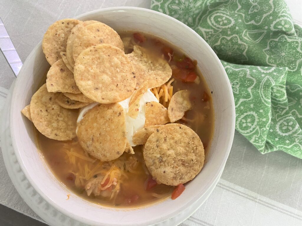 A bowl of chicken soup topped with round mini tortilla chips. sour cream and cheese with a green paisley napkin on a pretty table