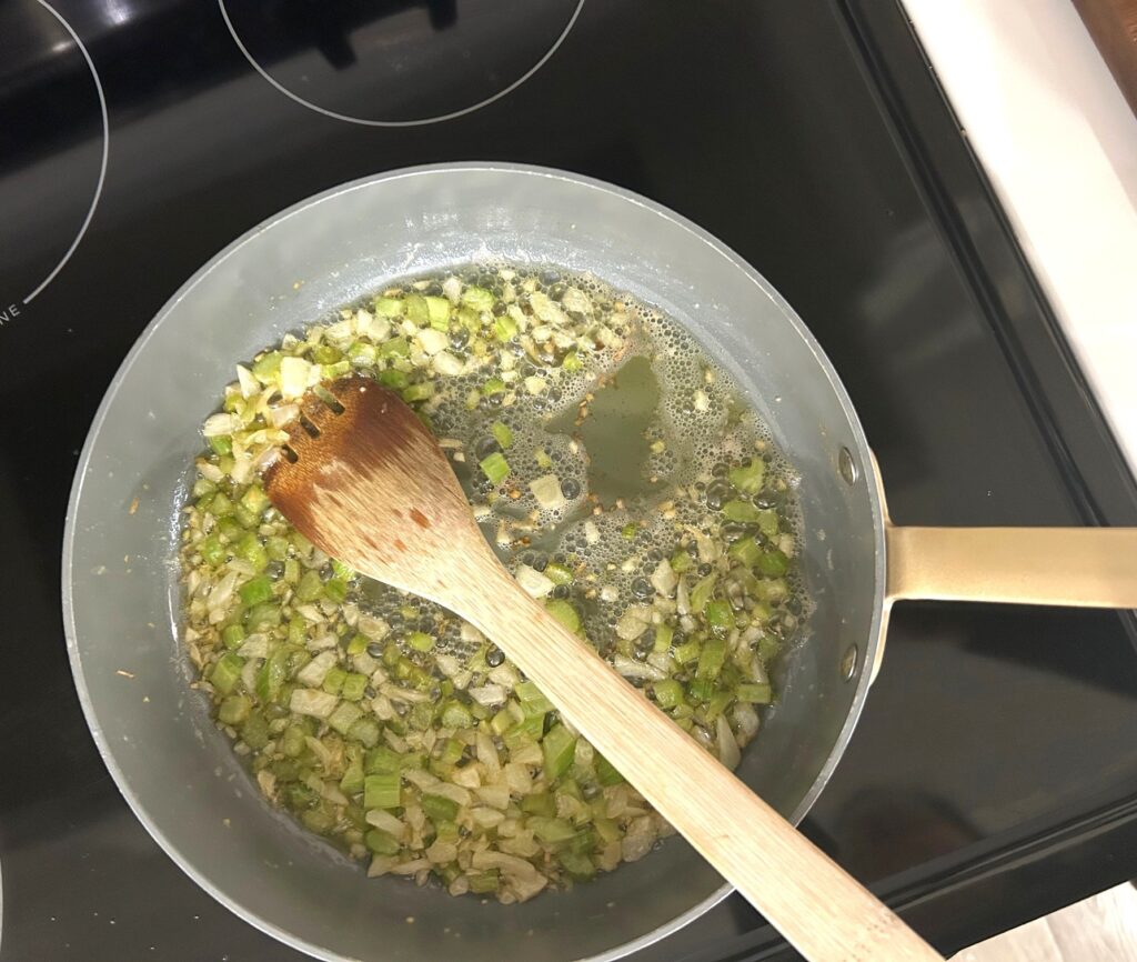 A pan on a black stovetop with onion, garlic, and celery being cooked in oil with a brown wooden spoon for stirring
