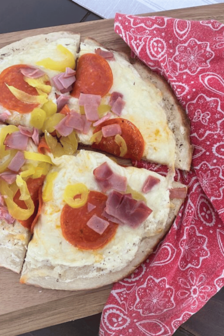 A personal sized homemade pizza topeed with cheese, pepperoni, ham and banana peppers on top of a red paisley cloth napkin