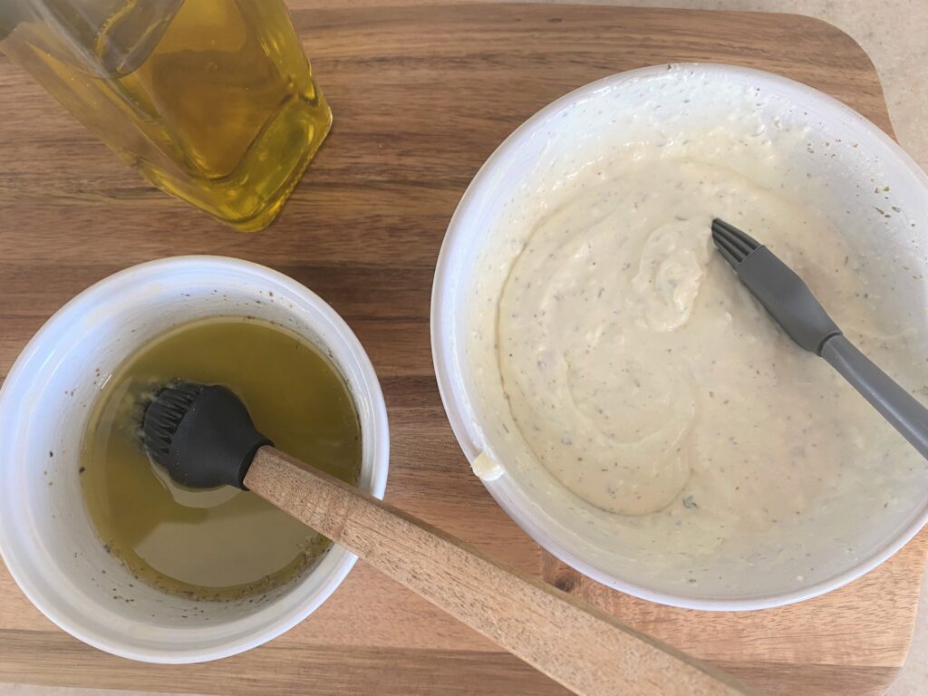 A wooden cutting board with white bowls of white pizza sauce and metled butter used to make homemade pizzas