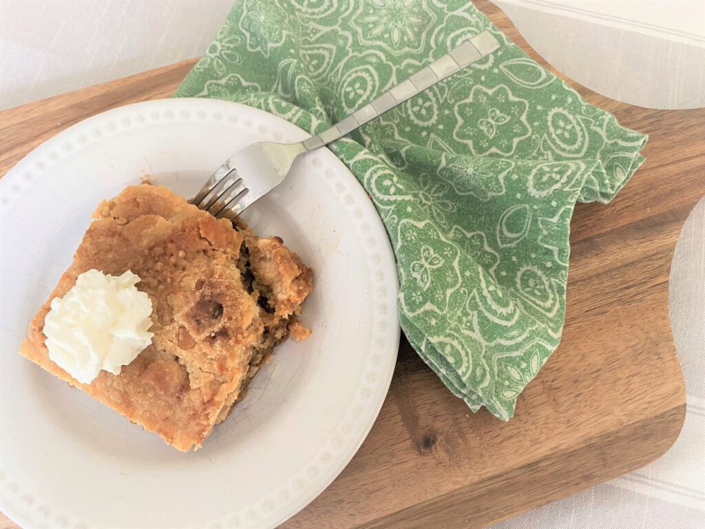 A wooden cutting board with a white place with a serving of pumpkin pie crunch bake topped with whipped topping and a green paisley towel next to it