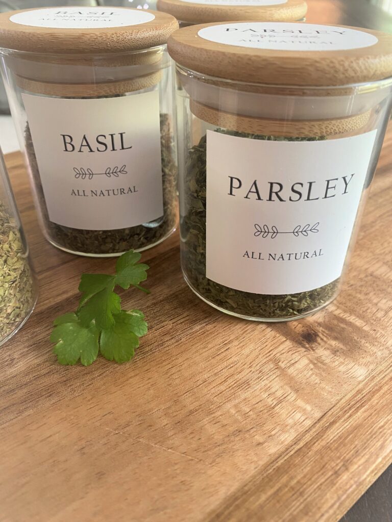 A close up picture of two glass spice jars with bamboo lids and pretty labs of parsley and basil with some parsley leaves on a wooden cutting board