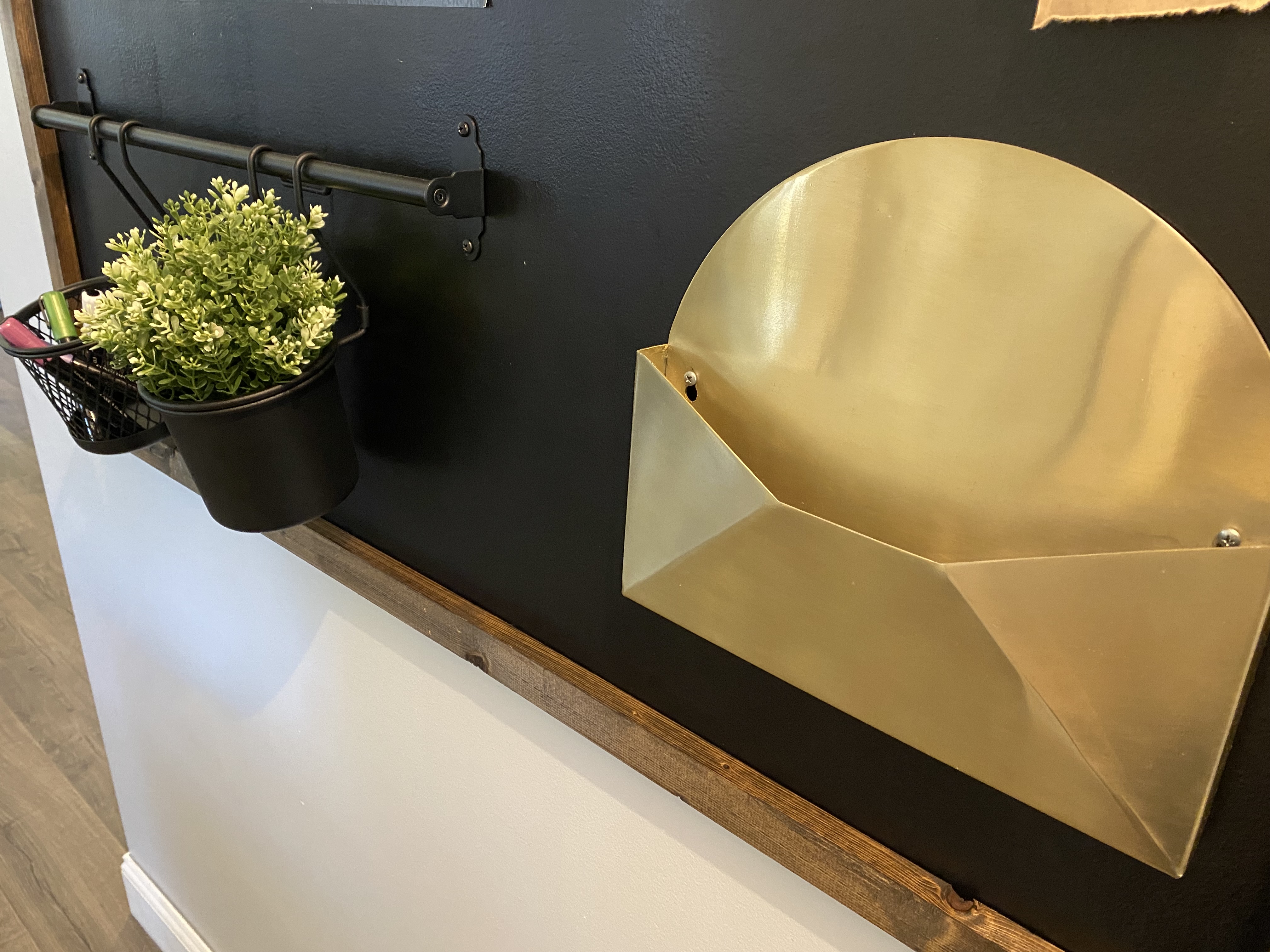 A gold wall file holder and a few black hanging baskets with a plant in one and chalkboard markers in the other to write on a wall chalkboard monthly calendar
