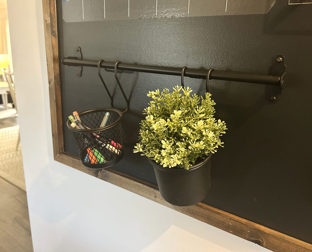 A close up picture of a black wall hanging rod with a hanging basekt of greenery and a hanging mash basket of chalk markers to write on a command center calendar