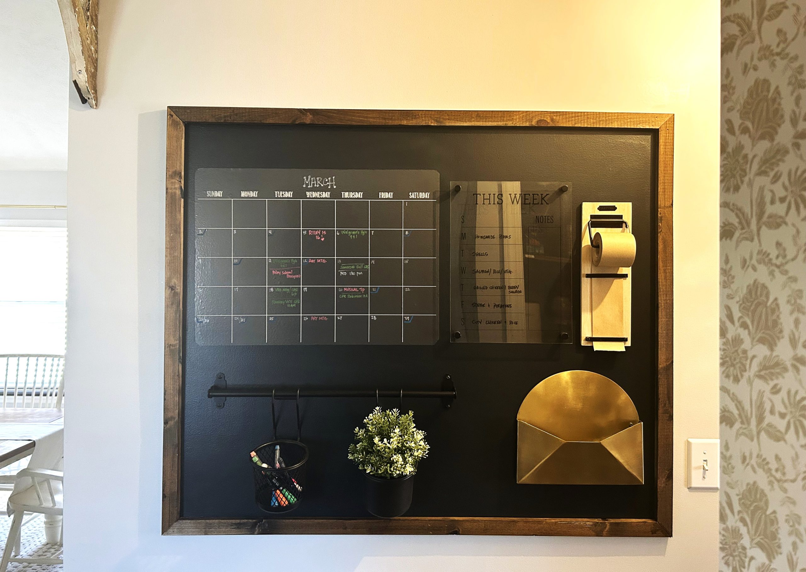 A wall command center on a hallway wall showing a wall calendar, menu board, golden paper file, hanging greenery basket and markers