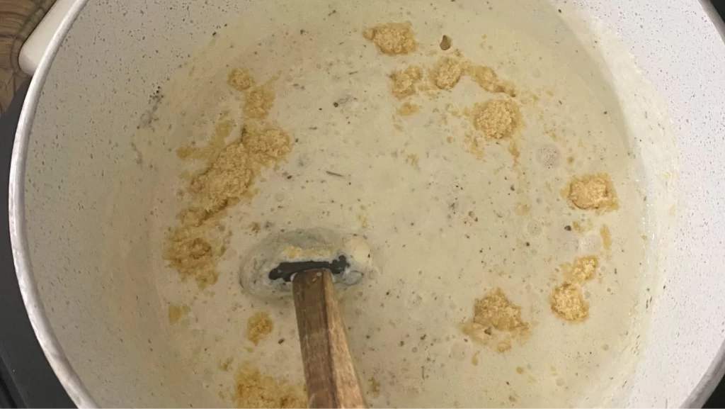 A white saucepan with wooden handles on a stovetop filled with homemade alfredo sauce with chunks of parmesan cheese being mixed in