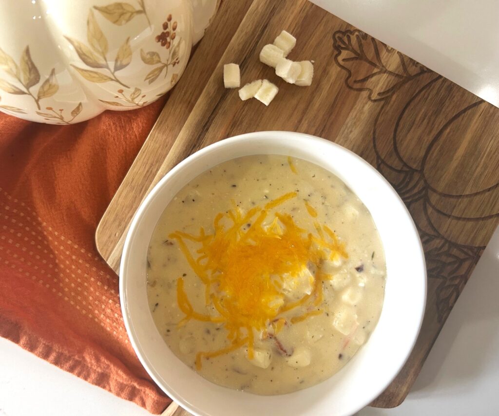 A wooden cutting board with a pumpkin on it and a bowl of homemade loaded potato soup topped with melted cheddar cheese