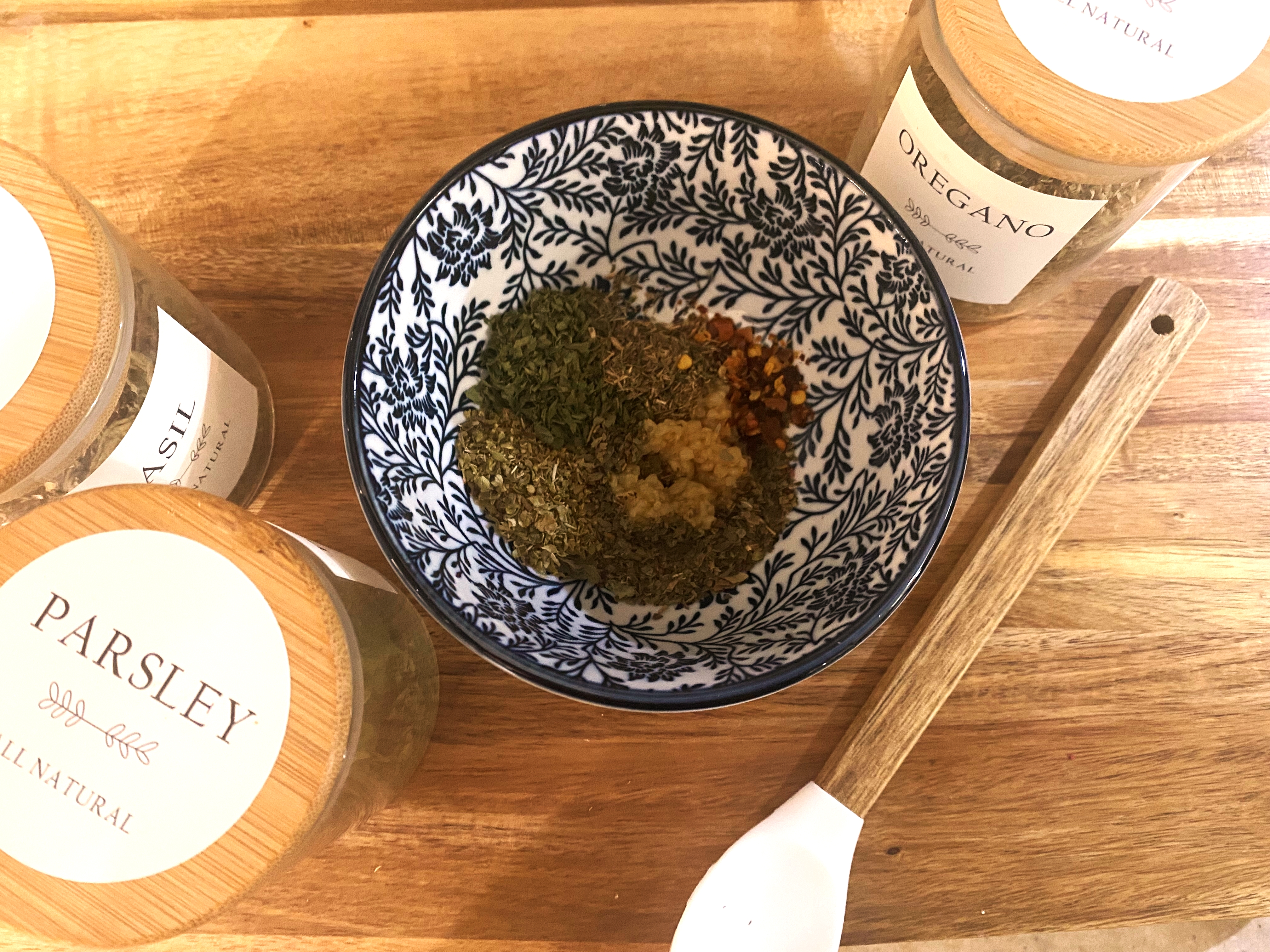 A wooden cutting board with a blue bowl of mixed spices next to labeled glass spice jars of parsley and oregano