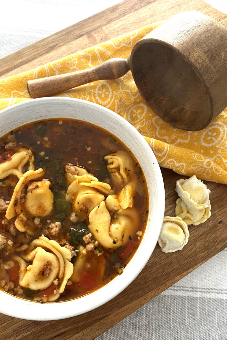 A bowl of sausage tortellini soup on a wooden cutting board with a wood ladle