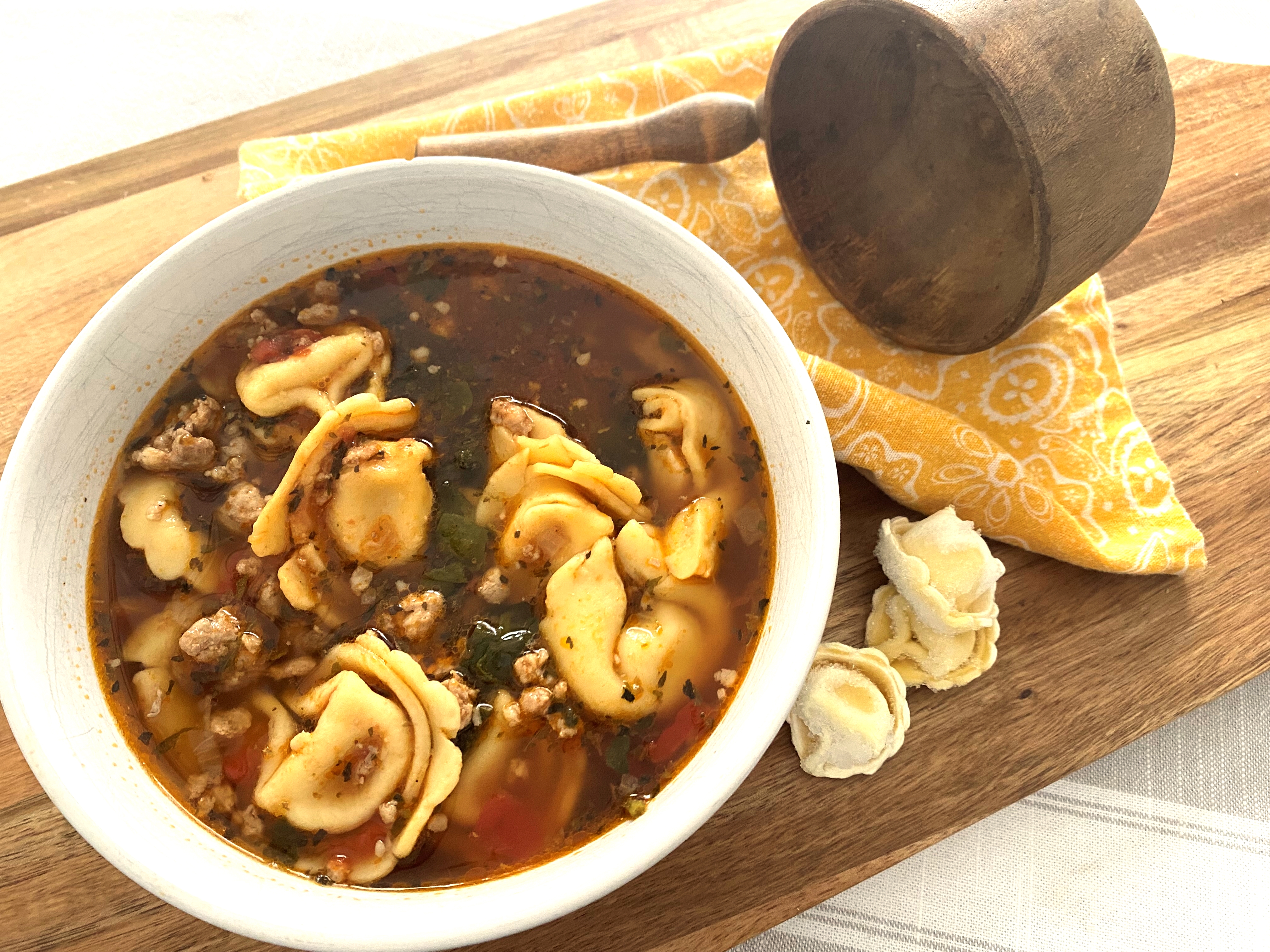 A bowl of sausage tortellini soup on a wooden cutting board with a wooden serving ladle