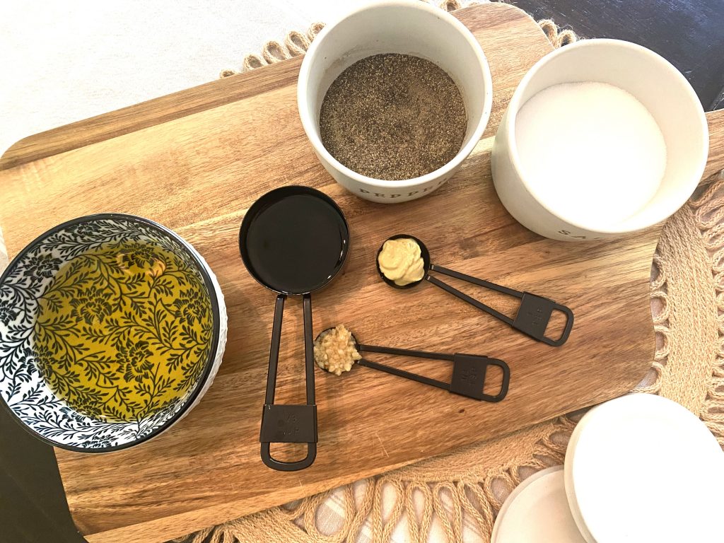 A wooden cutting board with bowls of premeasured olive oil, mustard, garlic, balsamic vinegar, and salt and pepper to make balsamic vinaigrette dressing