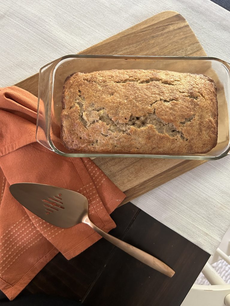 a wooden cutting board with a glass loaf pan with fresh baked banana bread with an orange tea towel and gold server