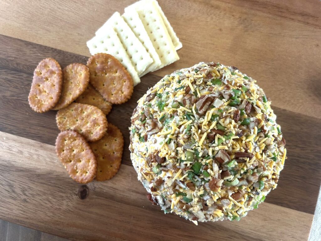 An overhead view of an assembled bacon and cheddar cheese ball, covered in bits of bacon, chopped pecans, green onion, and shredded cheese paired with crackers on a wooden cutting board