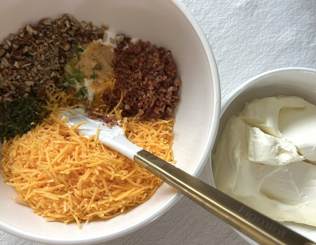 Two white mixing bowls of ingredients to make a homemade bacon cheddar cheese ball, one is softened cream cheese and the other is the remaining colorful ingredients of green onions, cheese, and spices