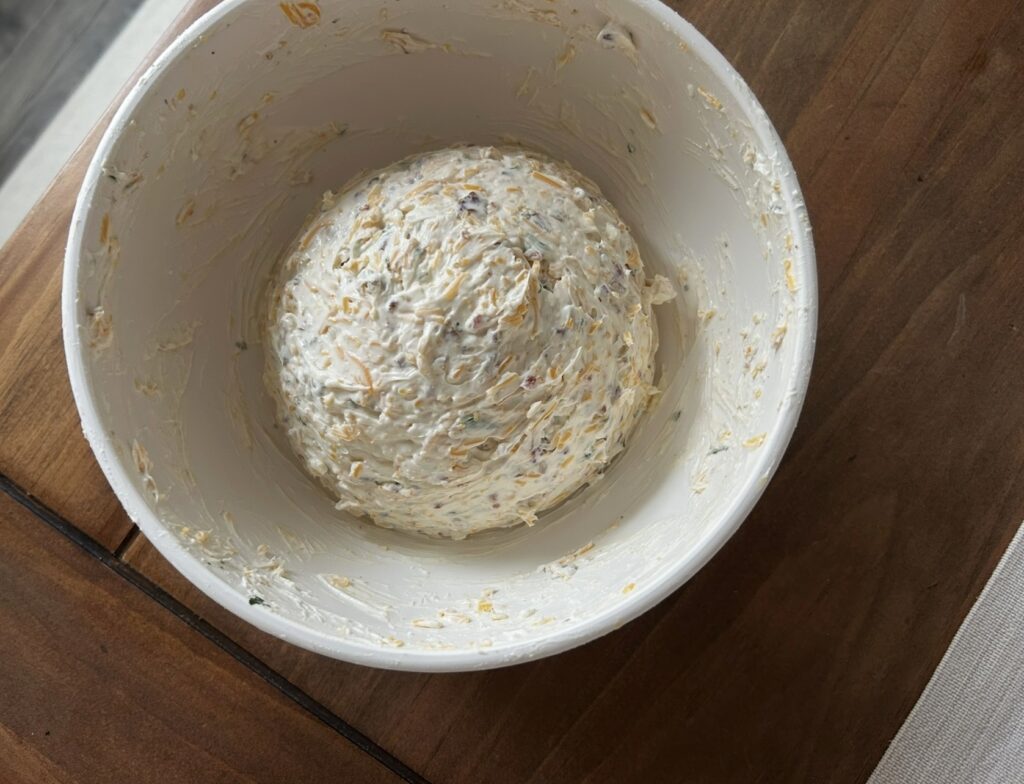 A white mixing bowl of a shaped cheddar bacon cheese ball not yet topped on a wooden table